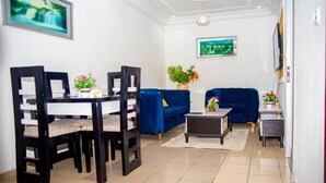 Una sala de estar con una mesa de comedor, sillas, un sofá azul, una pequeña mesa de centro y un televisor montado en la pared.
