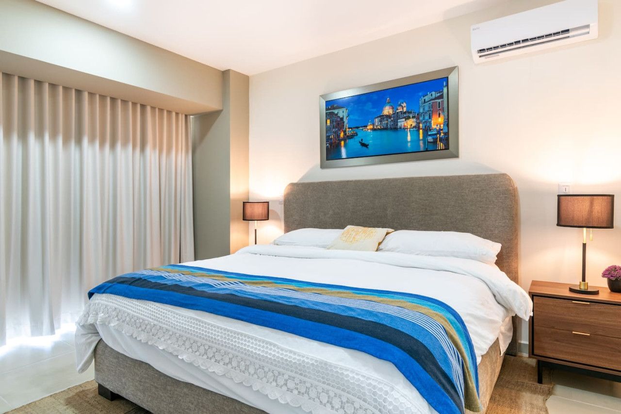 Una habitación de hotel con cama, mesita de noche, lámpara, un cuadro y una ventana con cortinas.