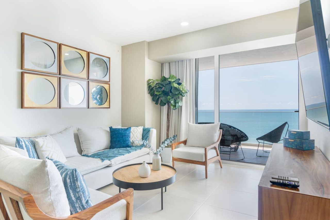 A modern living room with a sofa, chairs, a coffee table, and a TV. There is a balcony with a view of the sea.