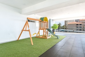 Zona de juegos infantiles con columpios y tobogán, ubicada en un balcón con vistas a un edificio de hotel.