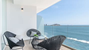 A balcony with wicker chairs and a small table, overlooking a clear blue sea and distant land.