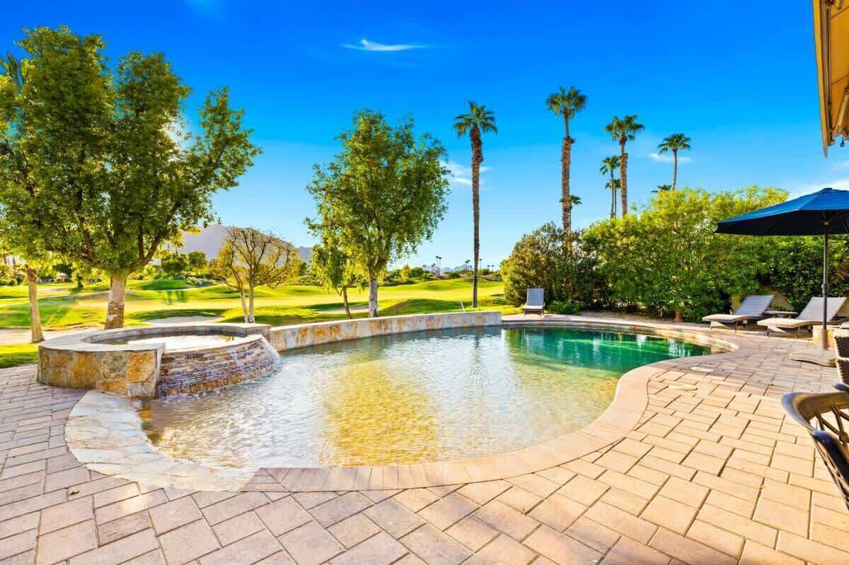 A swimming pool with a stone edge, surrounded by a brick patio, palm trees, and a view of a golf course.