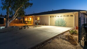 A residential house at dusk with a well-lit garage, a tree in the yard, and a clear driveway. - The Pink Agave: Retro Glam Views+Cold Plunge+Hot Tub+Gym+Dog&Festival Friendly (Yucca Valley)