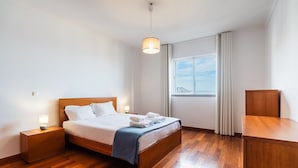 Et moderne hotelværelse med trægulv, en seng med hvide sengetøj, et natbord med lampe, et skrivebord og et vindue med gardiner.