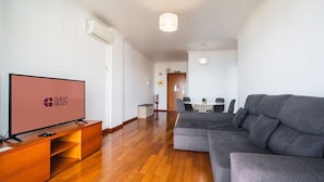 En moderne stue med et fladskærms-TV, en grå sofa, trægulv og et spiseområde med et bord og stole.