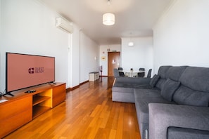 A modern living room with a flat-screen TV, grey sofa, wooden flooring, and a dining area with a table and chairs.