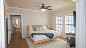 Une chambre avec un lit en bois, un ventilateur de plafond, une fenêtre avec des stores et une porte donnant sur une autre pièce.