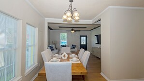 Un coin repas avec une table en bois dressée pour six personnes, des chaises à revêtement blanc, un lustre et un ventilateur de plafond.