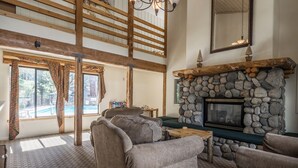 A spacious living room with a stone fireplace, large windows, and a high ceiling with wooden beams.