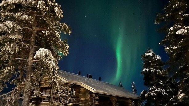 A log cabin with a snow-covered roof, surrounded by snow-laden trees, under a night sky with the aurora borealis. - Vacation home Naava by Interhome (Kittilä)