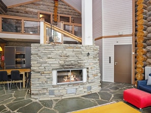 A stone fireplace with a fire, a dining area with chairs, a kitchen, and a staircase leading to an upper level.