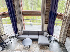 A modern interior with a grey sectional sofa, a round coffee table, and two side tables. Large windows with curtains provide a view of the outdoors.