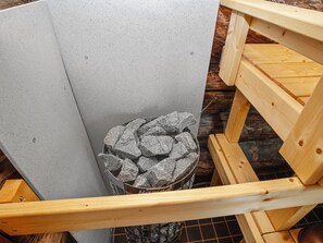 A corner of a room with a grey wall, a wooden bench, and a metal trash bin filled with rocks.