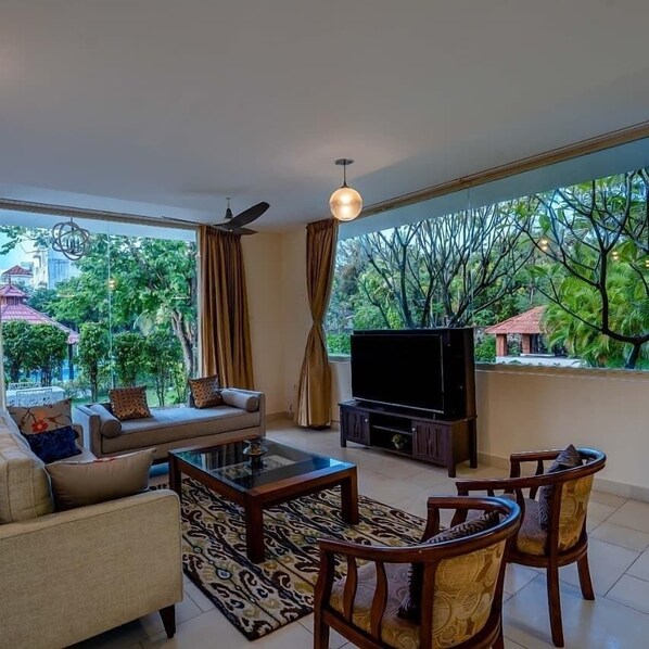 A living room with a sofa, coffee table, and chairs, a TV, and a large window with a view of greenery and buildings.