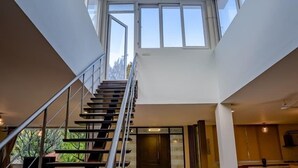 A modern interior with a staircase leading to a loft area, featuring large windows and a view of greenery outside. - Khushana Villa by Baywatch stayzz (Chennai)