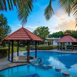 A swimming pool with a gazebo structure, surrounded by greenery and a clear sky.