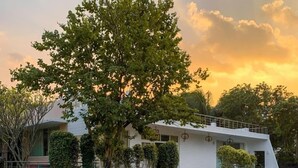 A modern building with a white exterior, surrounded by well-maintained hedges and trees. There are two lounge chairs with cushions and a table, suggesting a seating area. - Khushana Villa by Baywatch stayzz (Chennai)