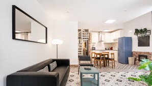 En stue med en sort sofa, et lille bord og en gulvlampe. Der er et tekøkken med et spisebord og stole, et blå køleskab og et flisegulv.