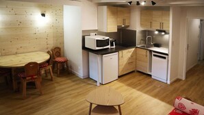 Eine kleine Küche mit Mikrowelle, Kaffeemaschine und Spüle. Ein Esstisch mit Stühlen und ein kleiner Hocker stehen bereit. Der Raum hat Holzböden und helle Wände.