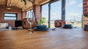 Un coin repas avec une table en bois dressée pour deux personnes, un mur en briques et une grande fenêtre donnant sur l’extérieur.