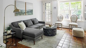 A living room with a grey sofa, a round coffee table, a floor lamp, and a large window with a view of greenery. - Enjoy Boston, Private Patio On Montgomery Park (Boston)