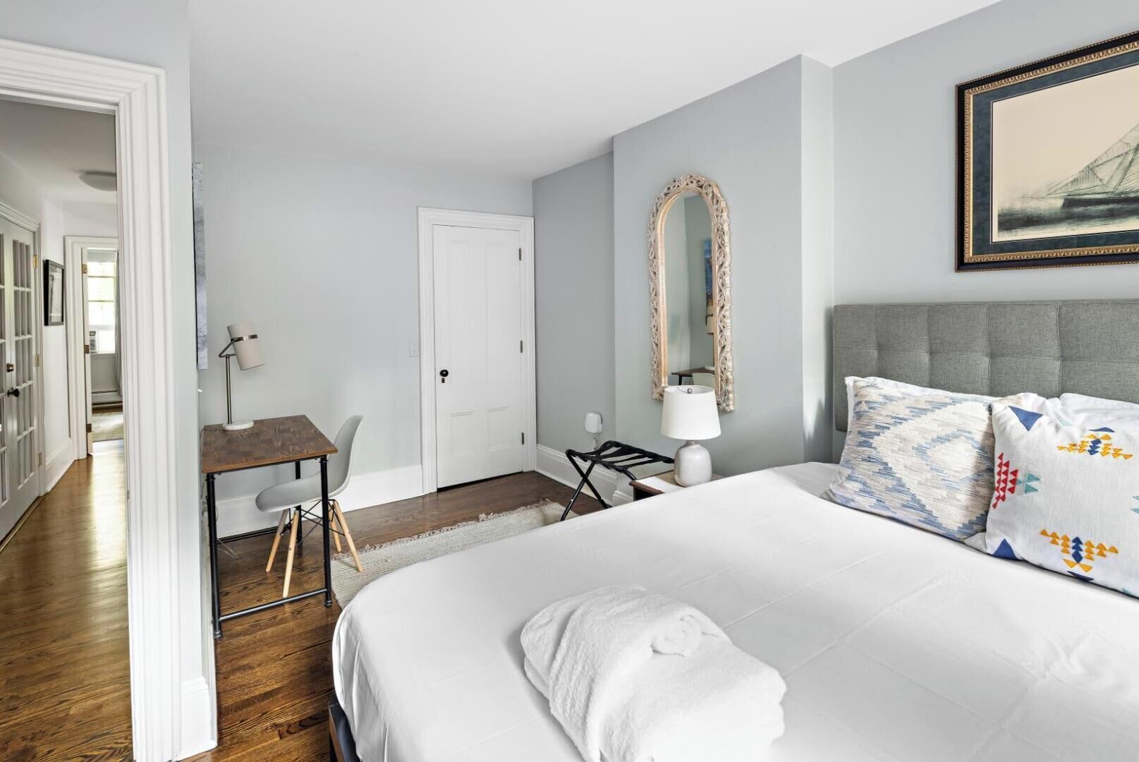 A hotel room with a bed, a desk, a chair, a mirror, and a painting on the wall.