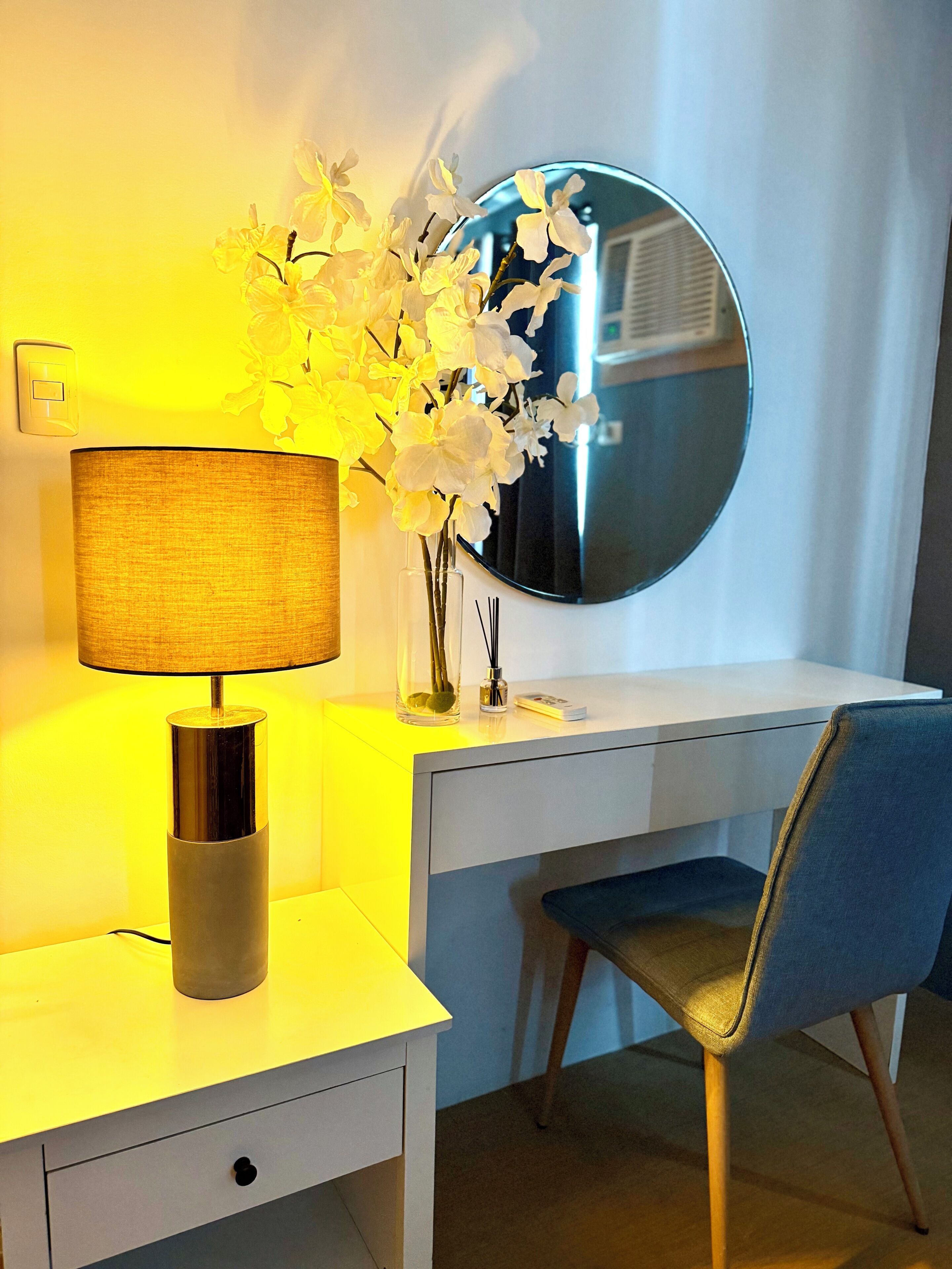 A modern room with a desk, chair, lamp, mirror, and a vase with flowers.