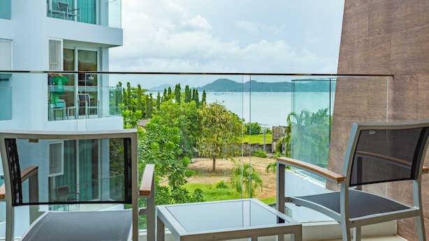 A balcony with a glass barrier, two chairs, and a view of a body of water and greenery. - Sea View Beach Dinning Beach Front c113 (Rawai)