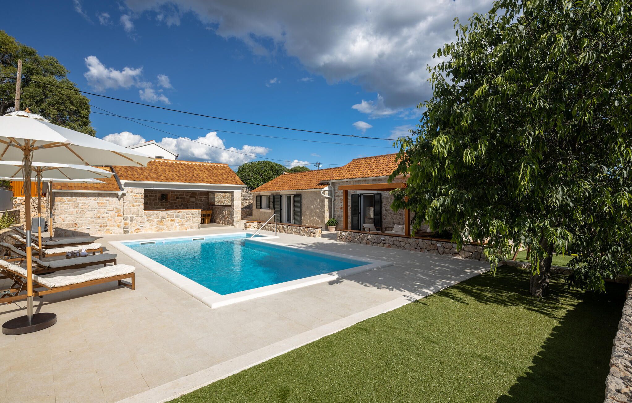 A swimming pool with lounge chairs and umbrellas, surrounded by stone buildings with tiled roofs.