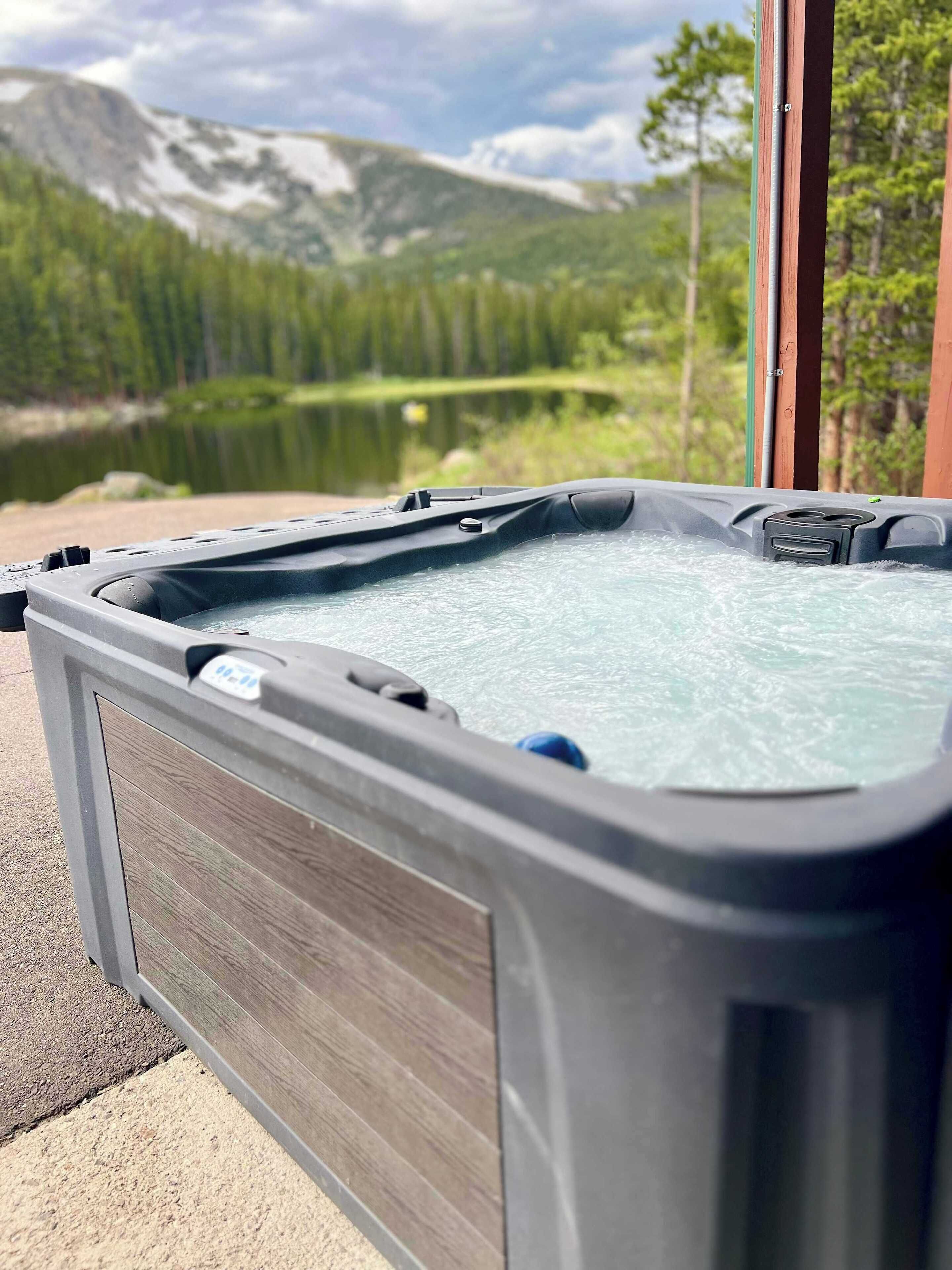 A hot tub with a wooden panel on the side, set against a backdrop of a lake and mountains.