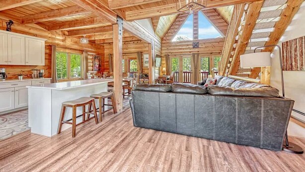 Ein geräumiges Blockhaus-Interieur mit einer großzügigen Sitzgruppe in der Mitte, Holzböden und einer hohen Decke mit freiliegenden Balken.