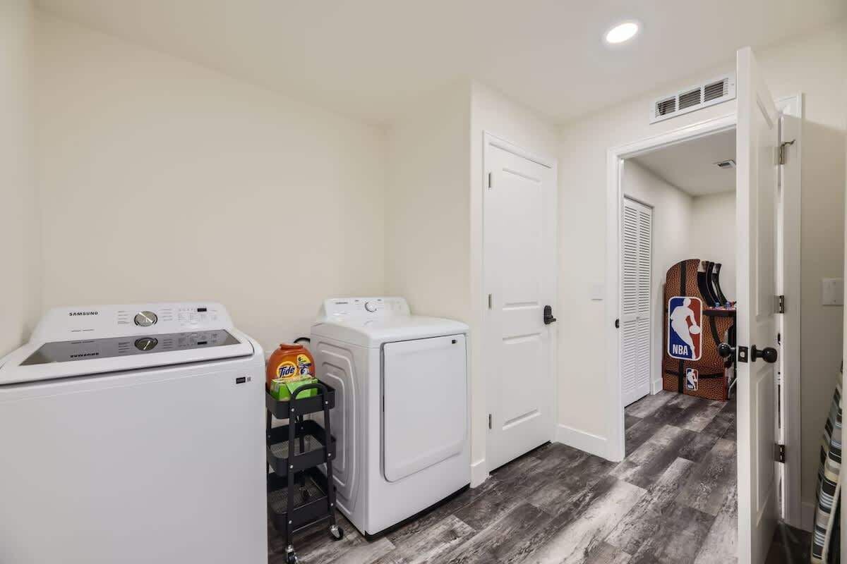 A laundry room with a Samsung washer and dryer, a cart with laundry detergent, and a door leading to another room with an NBA sign.