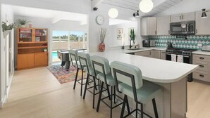 A modern kitchen with a central island, bar stools, a dining area, and a view of the outdoors.