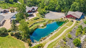 A large outdoor swimming pool surrounded by multiple buildings with sloped roofs, set in a lush, green landscape with hills in the background.