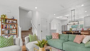 Een moderne woonkamer met een groene bank, witte fauteuils, een houten boekenkast en een trap naar een hoger gelegen verdieping.