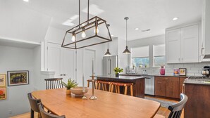 Een moderne keuken met houten kastjes, een centraal eiland, hangende lampen, en een eethoek met een houten tafel en stoelen.