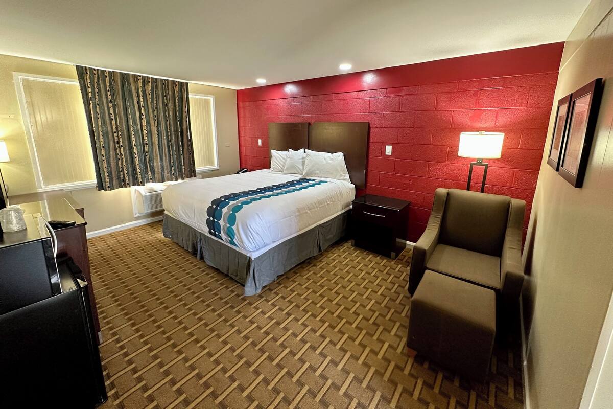 A hotel room with a large bed, a desk, a chair, a red brick accent wall, and a window with curtains.