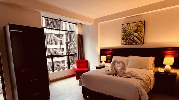 Una habitación de hotel con una cama, una silla roja, un televisor y un ventanal con vistas a edificios.