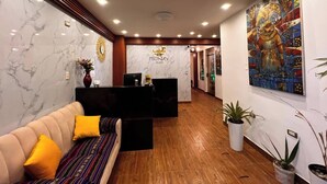 Zona de recepción de un hotel con pared de mármol, mostrador de recepción, sofá con cojines amarillos y un cuadro grande en la pared.