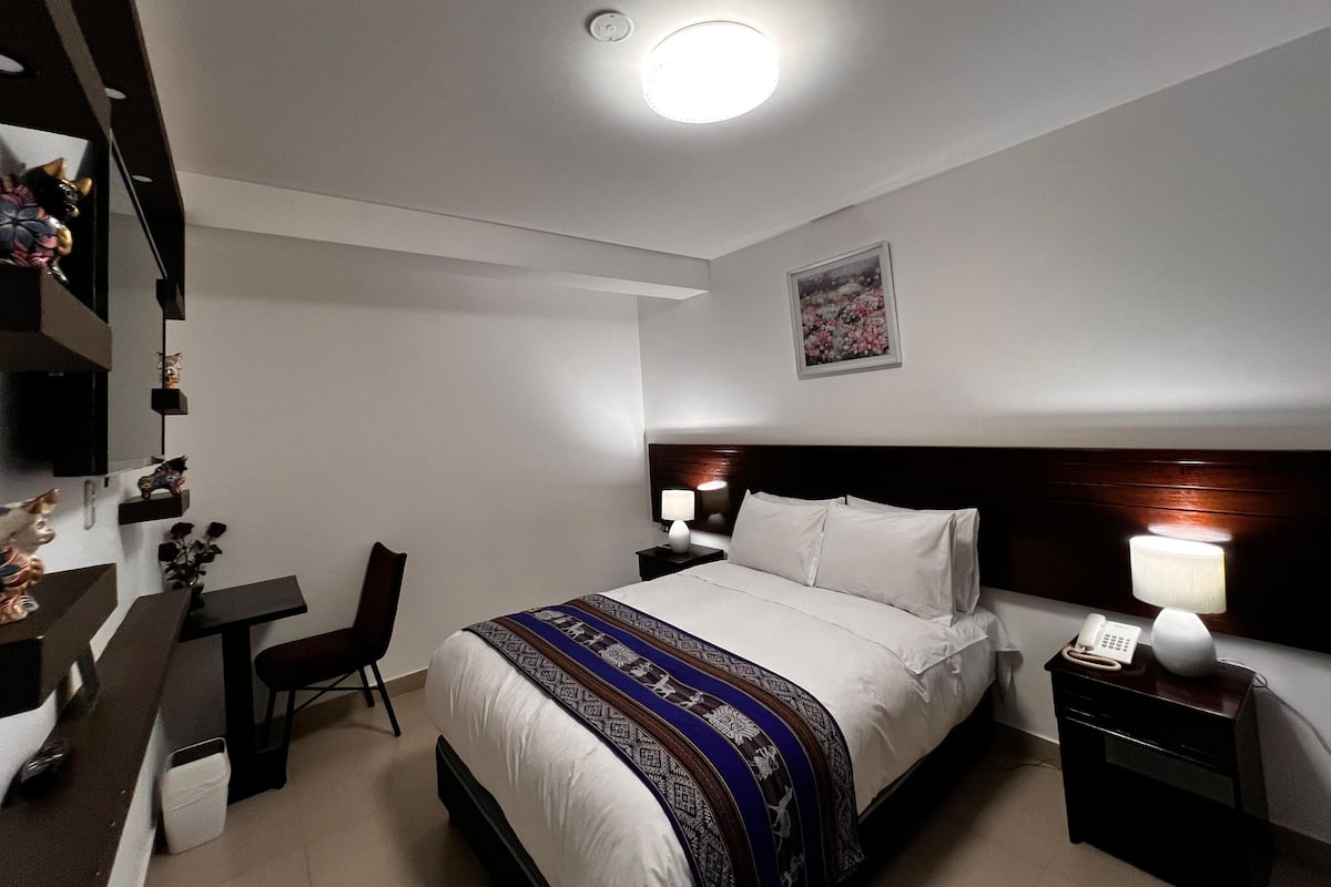 Habitación de hotel con cama, mesitas de noche, escritorio, silla y una pequeña estantería con objetos decorativos.