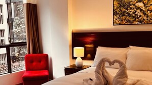 Una habitación de hotel con una cama, una silla roja, una mesita de noche con lámpara y una ventana con cortinas.