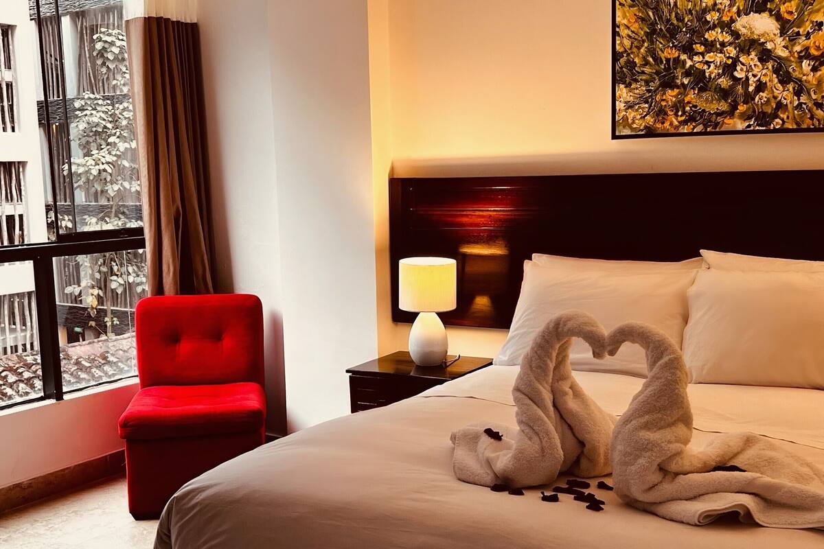 Una habitación de hotel con una cama, una silla roja, una mesita de noche con lámpara y una ventana con cortinas.