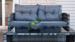 A wicker sofa with grey cushions and a potted plant on a balcony.
