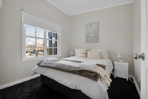 Un dormitorio con un ventanal grande, una cama con muchas almohadas, una mesita de noche y una obra de arte colgada en la pared.