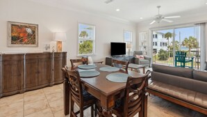 A spacious living area with a dining table, sofa, and TV. There are large windows and a balcony with outdoor furniture. - Amelia Sunrise - Oceanview, Bikes, & Beach Access (Fernandina Beach)