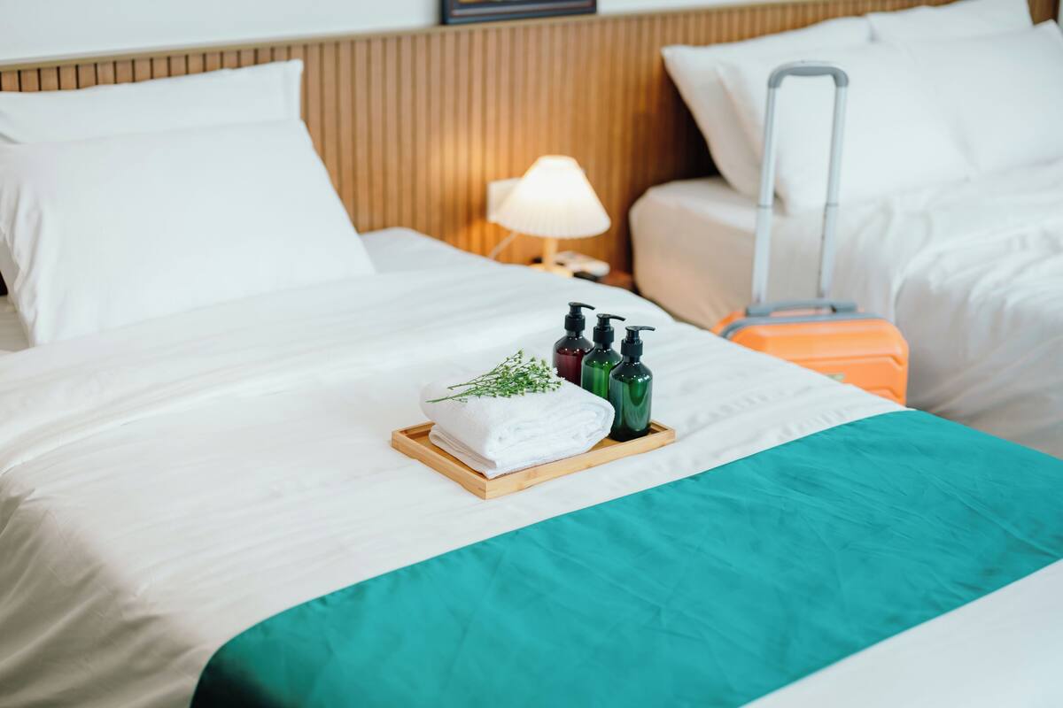 Un lit bien fait, recouvert d’une couverture turquoise, une petite table en bois avec des articles de toilette et une valise posée à côté.