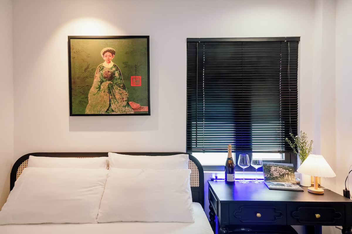 Une chambre d’hôtel avec un lit, une œuvre d’art encadrée au mur, une fenêtre avec des stores, un bureau avec une lampe et une bouteille de champagne.