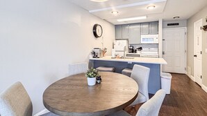 A modern kitchen with white cabinets, a central island, and a dining area with a round wooden table and grey upholstered chairs. - Inn Building Condo #107, ground level, pool view (Garden City)