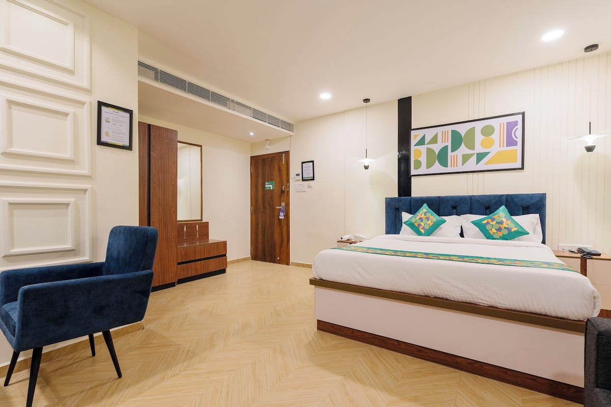 A hotel room with a bed, a blue chair, a wooden wardrobe, and a framed certificate on the wall.
