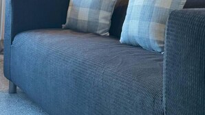 Un sofá gris oscuro con dos cojines a cuadros, una lámpara de pie alta y una ventana con cortinas.
